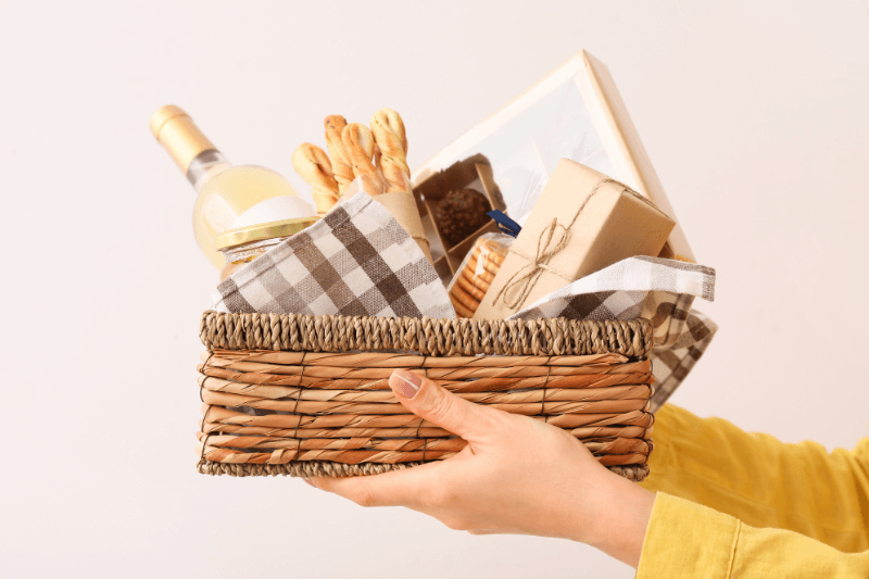 Hand holding wine gift basket with breadsticks, sweets, and wrapped boxes arranged neatly.