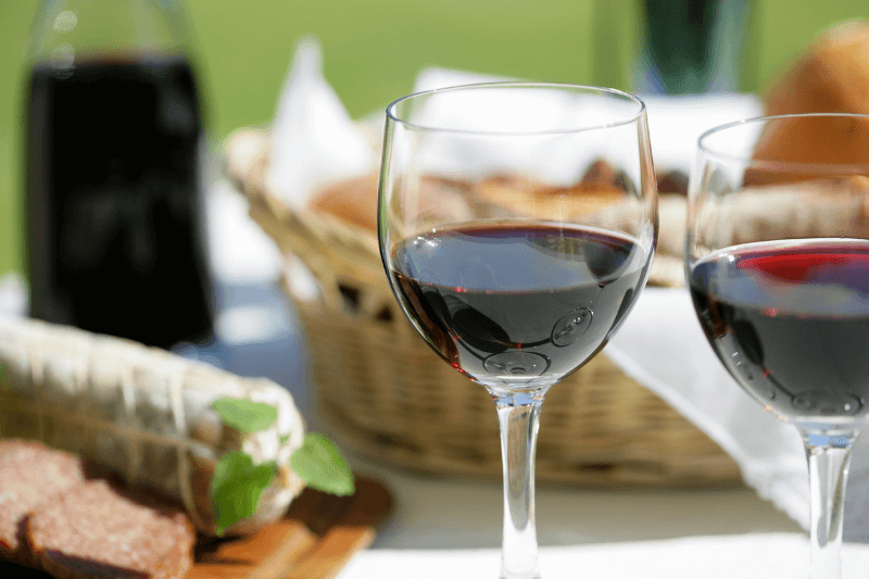 Two glasses of red wine with bread and picnic basket in outdoor setting.