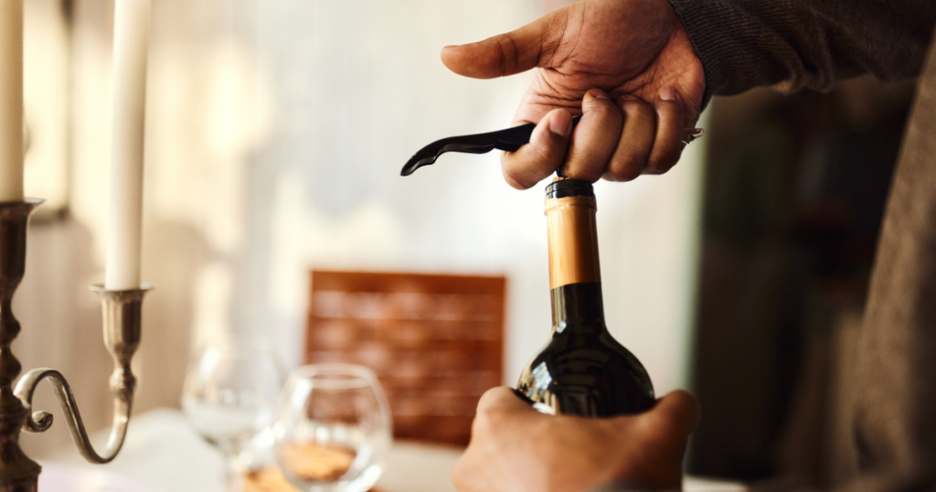 Person's hand using a traditional corkscrew with wooden handle to open a wine bottle at a dining table setting