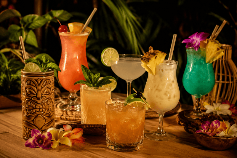 Tiki cocktails with tropical fruit garnishes on a wooden table with palm leaves in the background