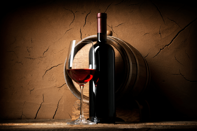 Red wine bottle and glass displayed beside a wooden barrel in a wine cellar